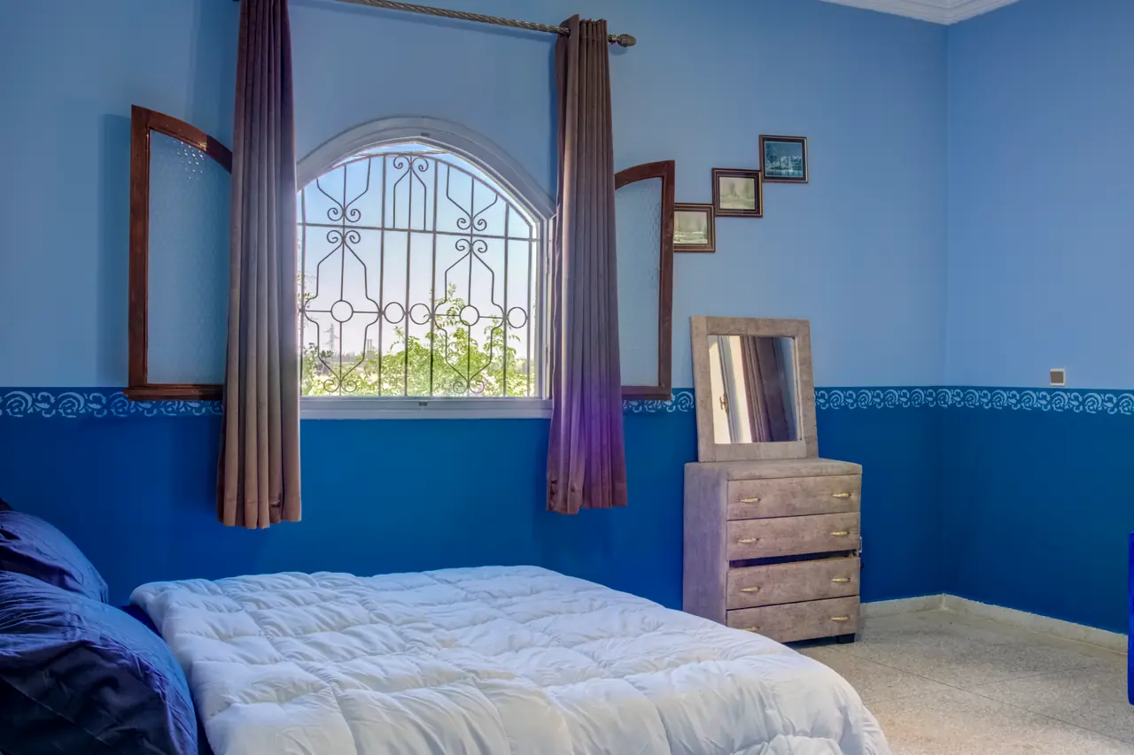 Chambre traditionnelle aux murs bleus avec fenêtre cintrée en fer forgé marocain