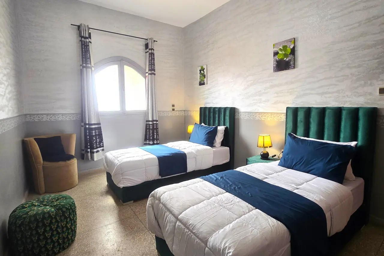 Chambre avec deux lits simples, têtes de lit en velours vert et fauteuil de lecture