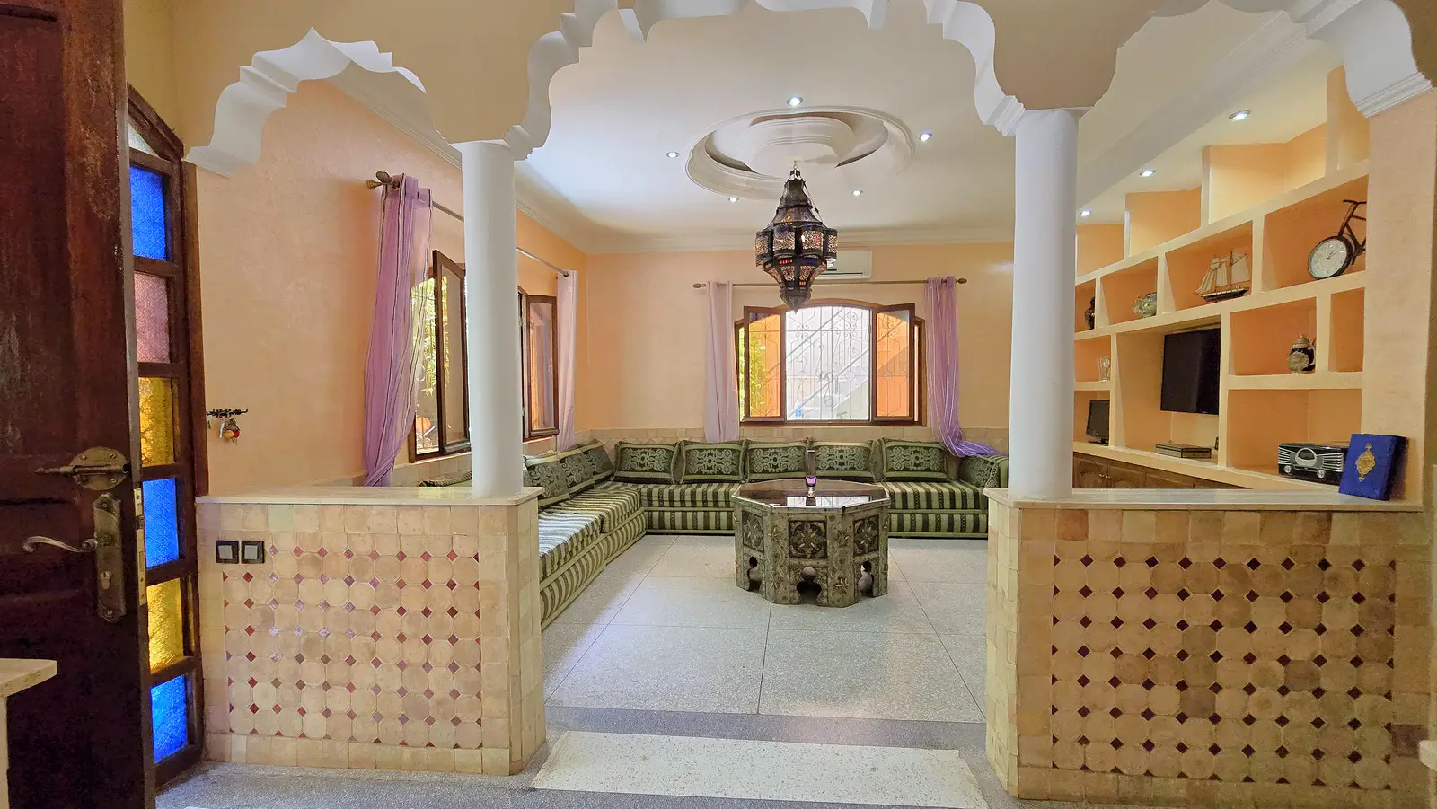 Large Moroccan salon — traditional U-shaped seating, carved octagonal table, arches and stained-glass door